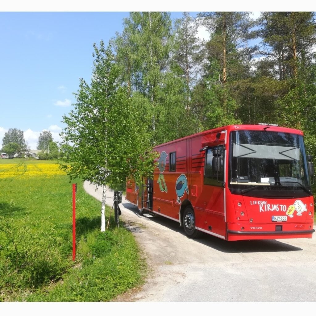Kuvassa on kirjaustoauto pysäköitynä tien risteykseen. Ympärillä näkyy puita ja kukkia täynnä oleva pelto.