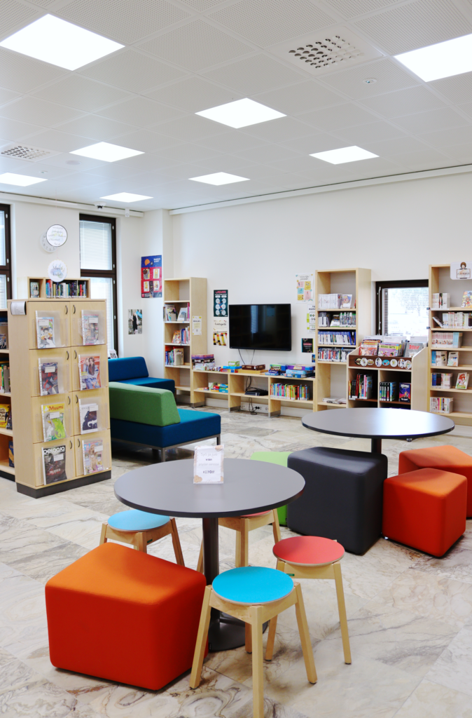 Kuvassa on Lieksan kirjaston lasten- ja nuortenosastoa. Kuvassa etualalla on kaksi pöytäryhmää. Taaempana on kirja- ja pelihyllyjä ja televisio.