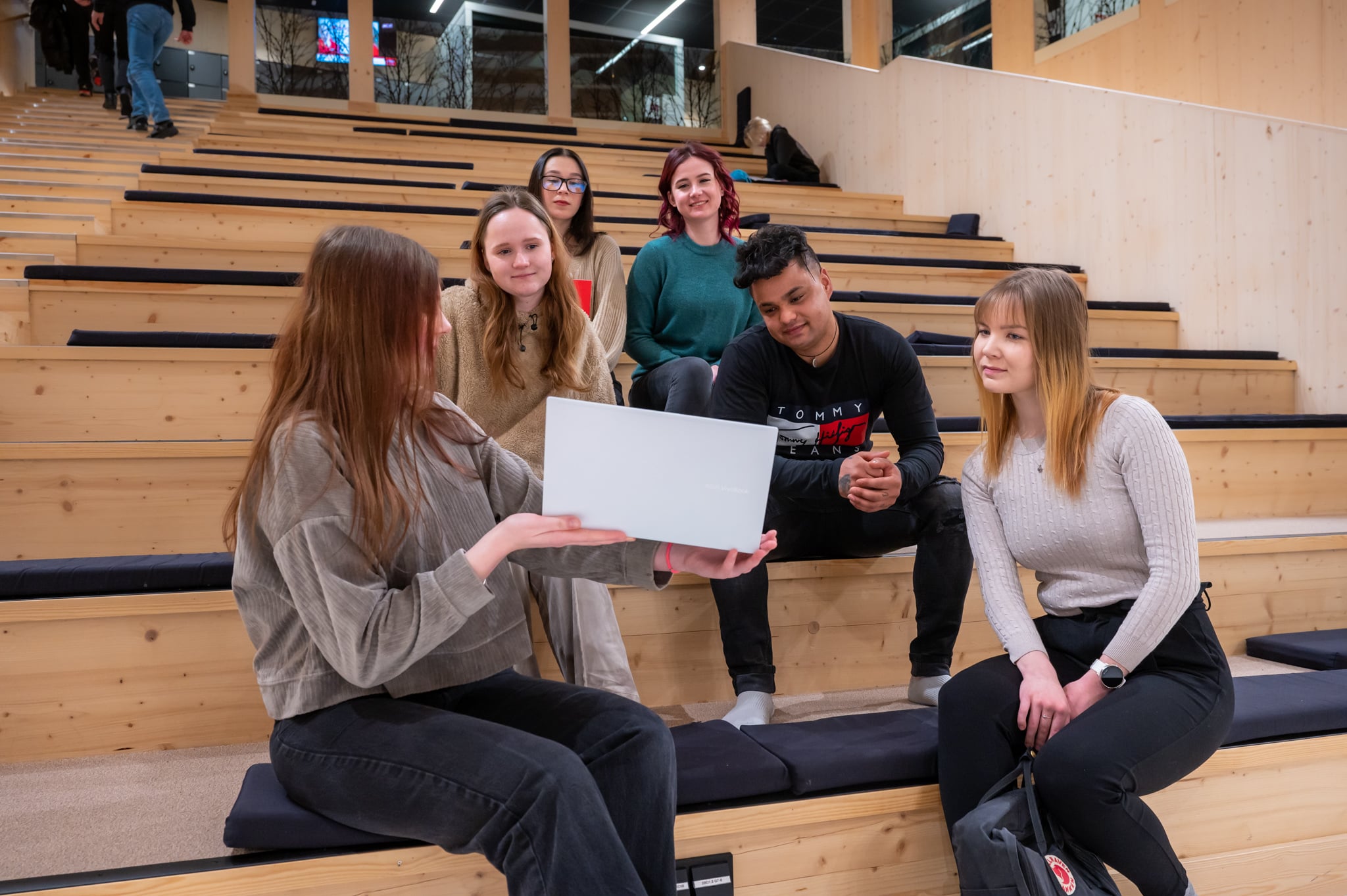 Nuoria ihmisiä istumassa rappusilla. Edessä oleva nuori nostaa kannettavaa tietokonetta, jonka näyttöä kaikki katsovat.