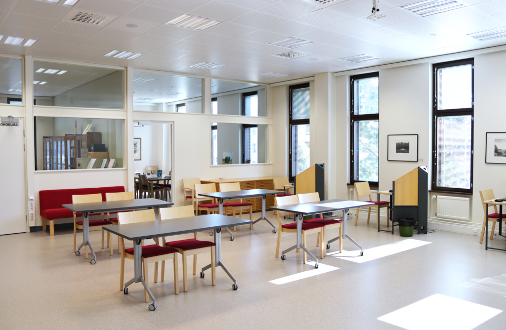 Kuvassa on Lieksan kirjaston matineatila. Tilassa on neljä pöytää ja tuoleja. Taustalla näkyvät kirjaston kotiseutukokoelmahuone ja mummolahuone.