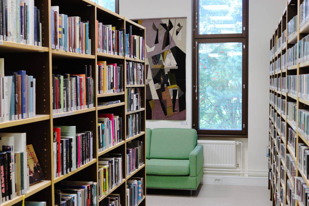 Kuvassa on kirjaston musiikkiosasto. Molemmilla sivuilla näkyy kirja- ja levyhyllyjä, keskellä sohva ja ikkuna. Ikkunan vieressä seinällä on tekstiilitaideteos.