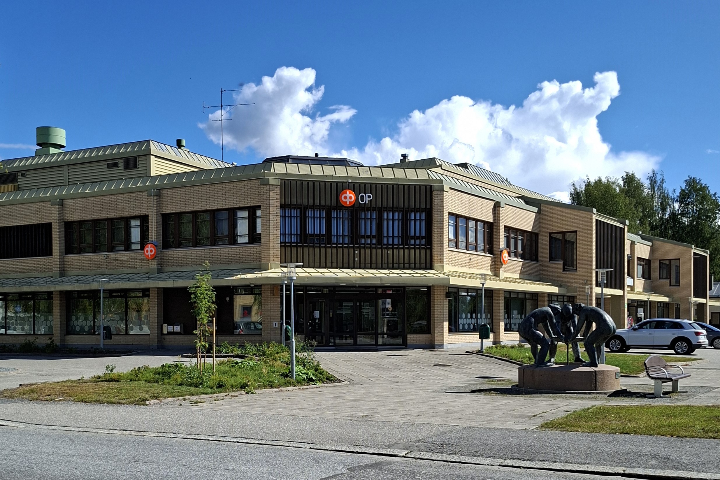 Kaksikerroksinen liikekiinteistö. Etualalla patsas, jossa kolme hahmoa kumartuneena kannattelee yhdessä astiaa.