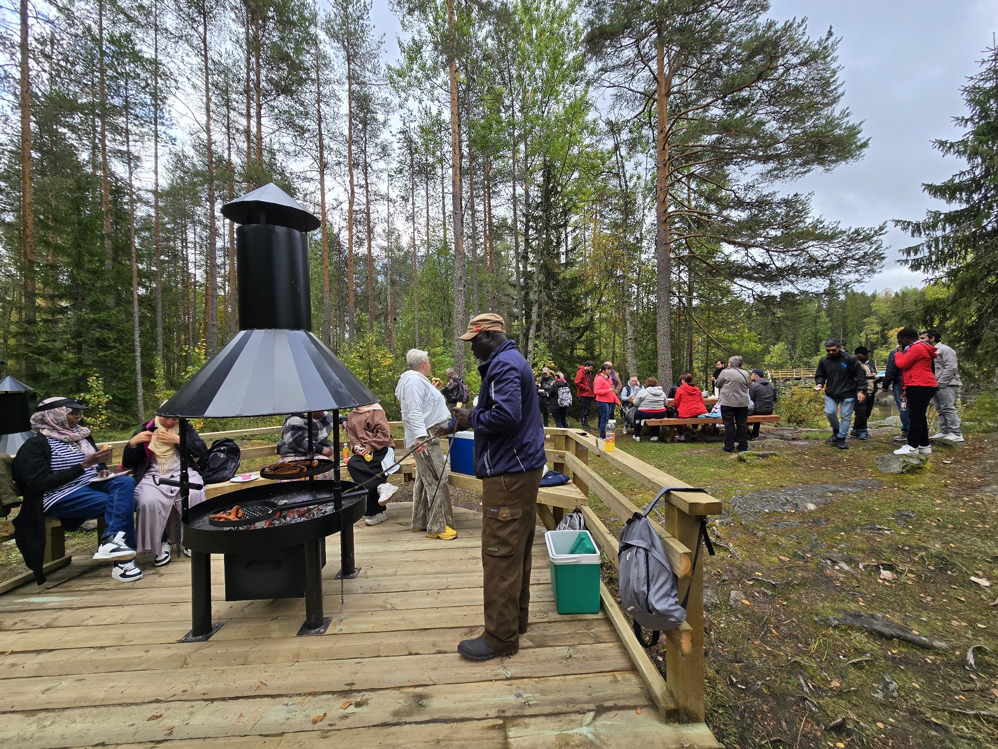 Monen eri kansallisuuden ihmisiä metsässä retkellä. Osa paistaa makkaraa grillissä.